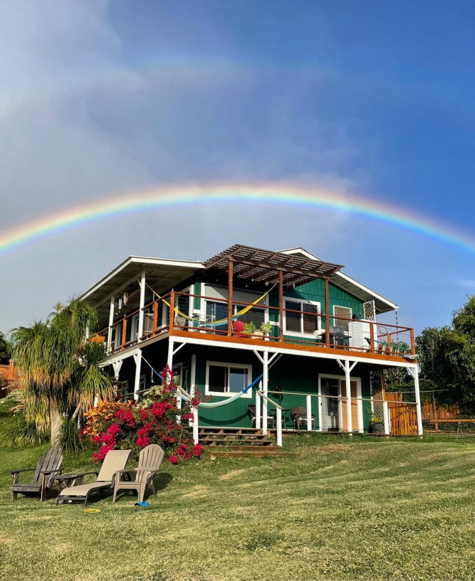 A two-story green house with white trim is framed by vibrant pink bougainvillea and a gentle rainbow in the sky. A wooden deck wraps around the house, featuring lounge chairs positioned on the lawn, providing a relaxing outdoor space.