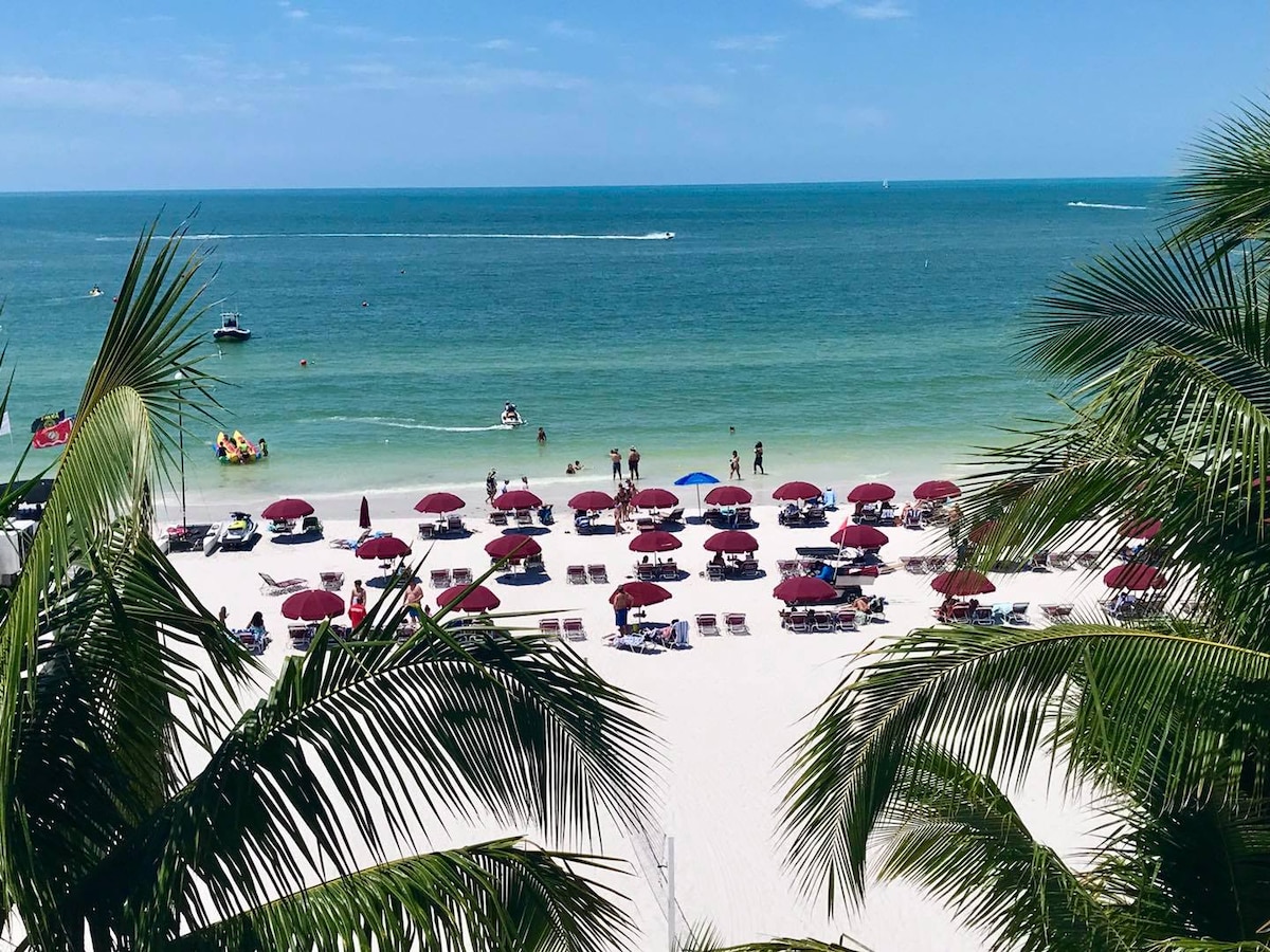 A sandy beach is lined with colorful umbrellas and sun loungers, providing ample space for relaxation. Gentle waves can be seen lapping at the shore, while people engage in beach activities. Lush palm fronds frame the view, adding a touch of greenery to the seaside scene.