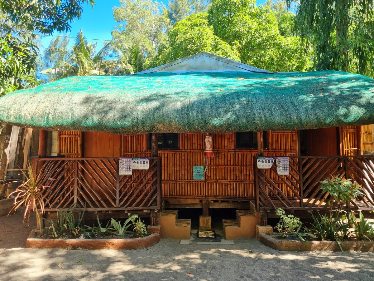 The rustic cottage features a thatched roof and wooden walls, surrounded by tropical vegetation. The front porch is adorned with potted plants, and notices are displayed on the wooden slats. The structure is situated in a sandy area, providing easy access to the beach.