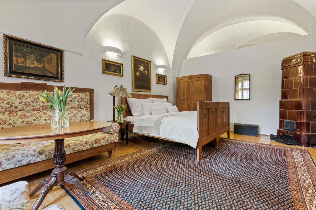 A spacious bedroom features a large bed with crisp linens and a comfortable seating area comprised of a floral-patterned sofa and a wooden table. Illuminated by soft lighting, the room is adorned with artwork and a decorative tiled stove, enhancing the historic ambiance.