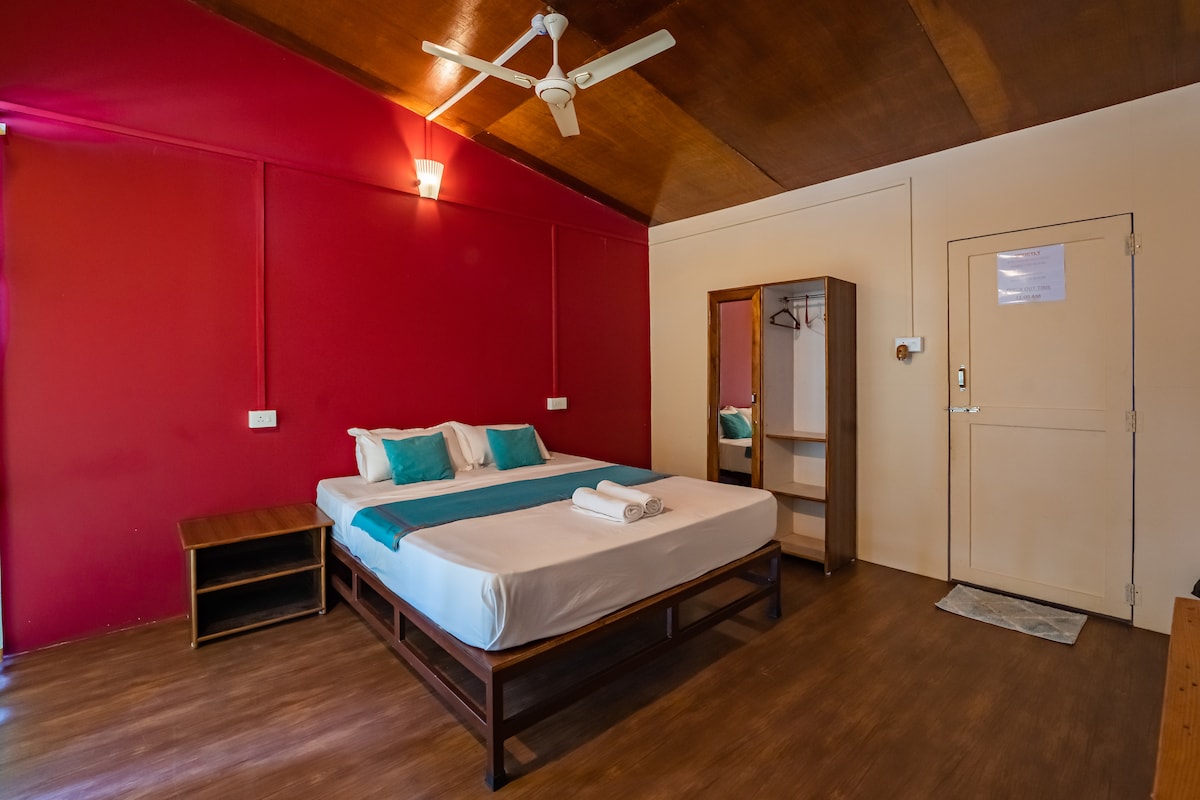 A well-appointed sleeping area features a spacious bed with light blue accent pillows. The textured wall is painted in a warm red hue, complemented by wooden ceiling panels. A mirror and open closet sit adjacent to the bed, with a door leading outside on the far wall.
