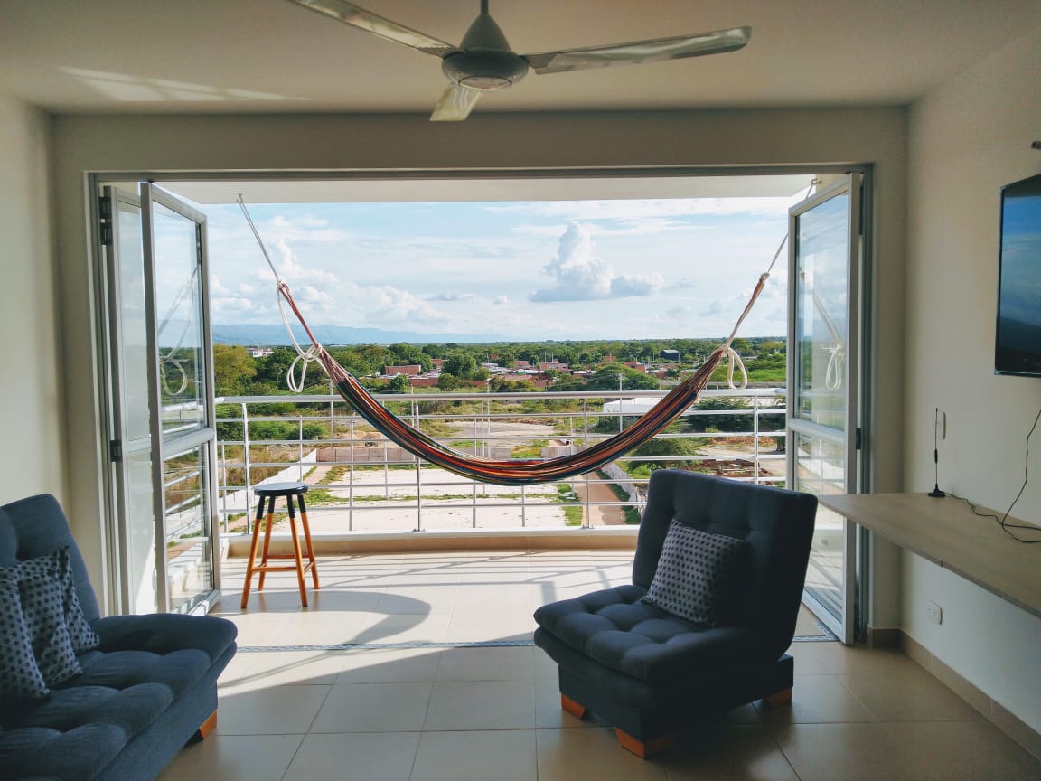An open living space features large glass doors that lead to a balcony with a scenic view. A colorful hammock hangs between the doors, inviting relaxation. Two comfortable armchairs and a side table complement the area, which is well-lit by natural light.