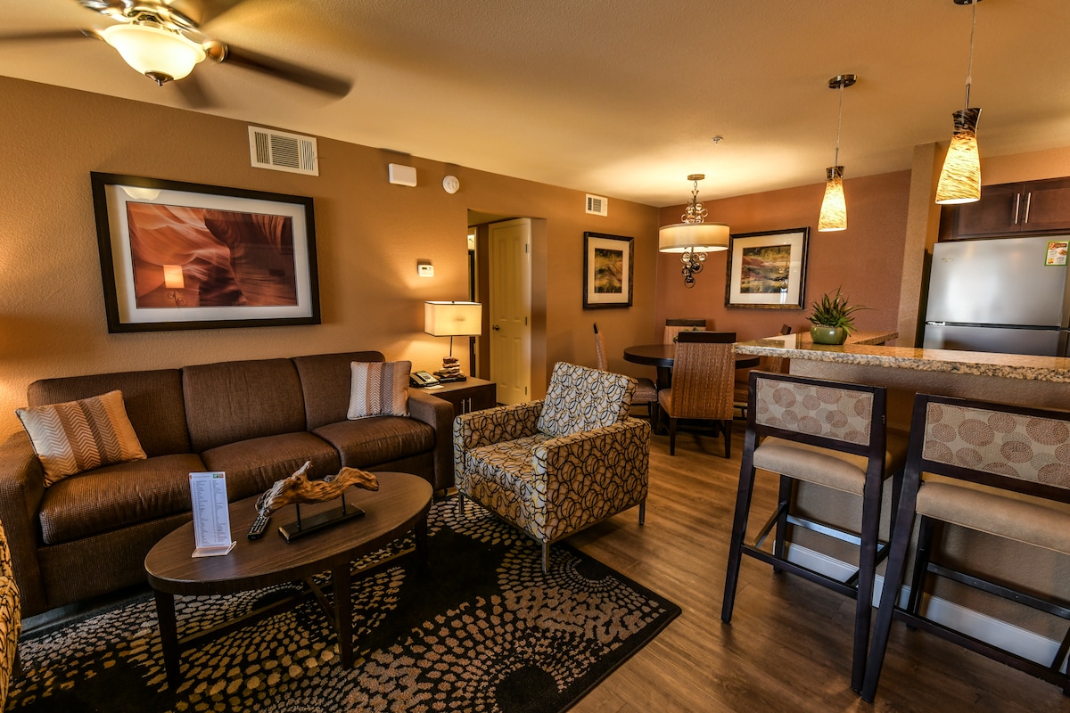 An inviting living area features a comfortable sofa adorned with pillows, accompanied by accent chairs. A wooden coffee table centers the space, with a patterned area rug beneath. The room is illuminated by warm lighting, and an open kitchen area with modern bar stools is visible in the background.
