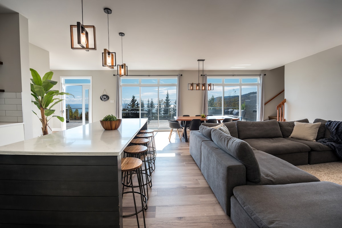 An open living area showcases a spacious sofa facing the dining area, which features a large table with surrounding chairs. Natural light floods the space through large windows, revealing scenic views. A kitchen island with bar stools offers additional seating and convenience.