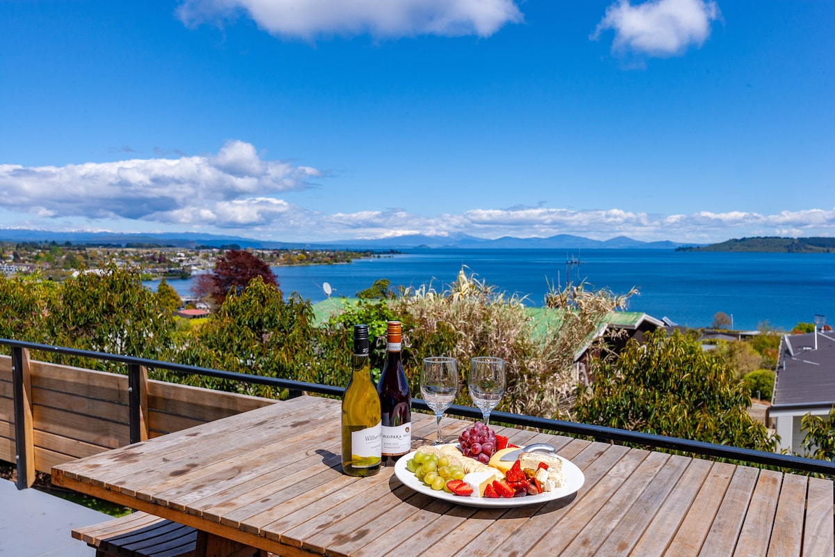 A dining setting is positioned on a wooden deck, featuring a round platter with assorted cheeses, fruits, and wine bottles. Scenic views of the lake and mountains are visible in the background under a clear blue sky, creating a relaxing atmosphere.
