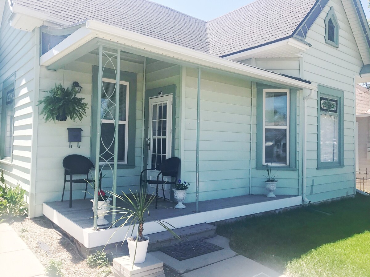 A charming front porch features two black chairs positioned beside a small table. Potted plants add greenery to the space, while a decorative railing frames the entrance. The light blue exterior of the cottage enhances the welcoming atmosphere.