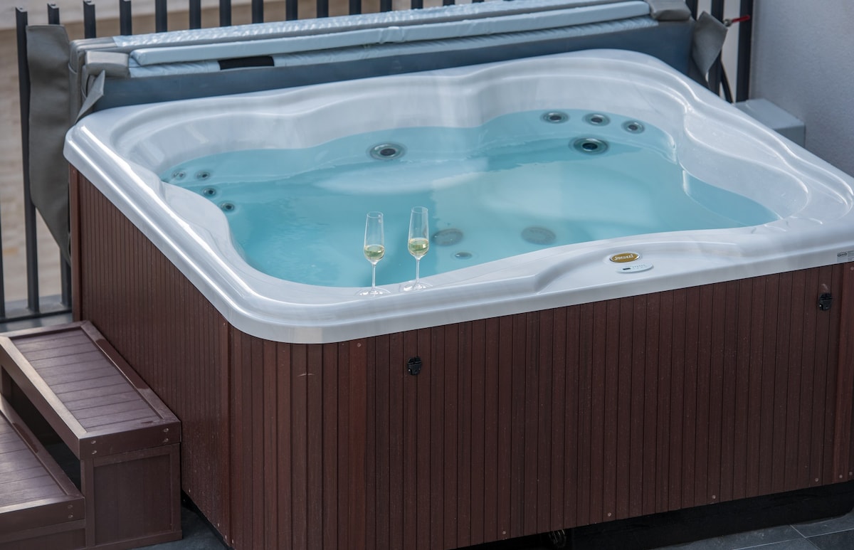 A hot tub is situated on a private terrace, featuring clear water and jets. Two glasses sit elegantly on the edge. The setting offers an inviting space for relaxation, framed by a minimalist wooden enclosure.