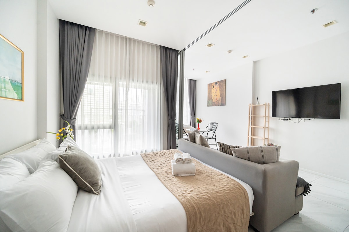 A spacious bedroom area features a comfortable bed adorned with neutral-toned bedding. A large window allows natural light to fill the space, complemented by sheer curtains. A cozy sofa sits nearby, accompanied by decorative pillows, while modern art decorates the wall opposite the bed.