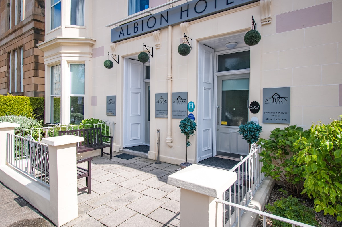 The entrance of the Albion Hotel features a dual-door entry flanked by green foliage and decorative hanging planters. A bench is positioned on a tiled patio, bordered by a low white fence. Clear signage indicates the hotel's name and its welcoming atmosphere.