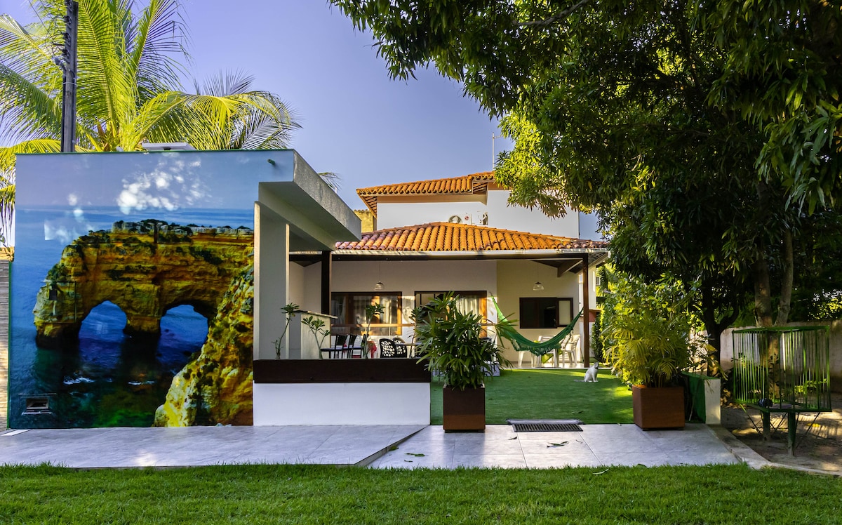 A well-maintained exterior is highlighted by a vibrant mural featuring a coastal rock formation. A spacious green lawn is complemented by palm trees and outdoor seating. The house showcases a traditional tiled roof and an inviting patio area, creating a welcoming outdoor space.