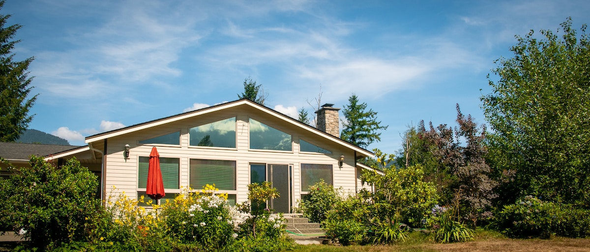 The exterior of a spacious riverside home is visible, adorned with vibrant greenery and flowering plants. A red umbrella provides shade over the patio steps, inviting outdoor relaxation. Large windows reflect natural light, creating a connection between the indoor and outdoor environments.