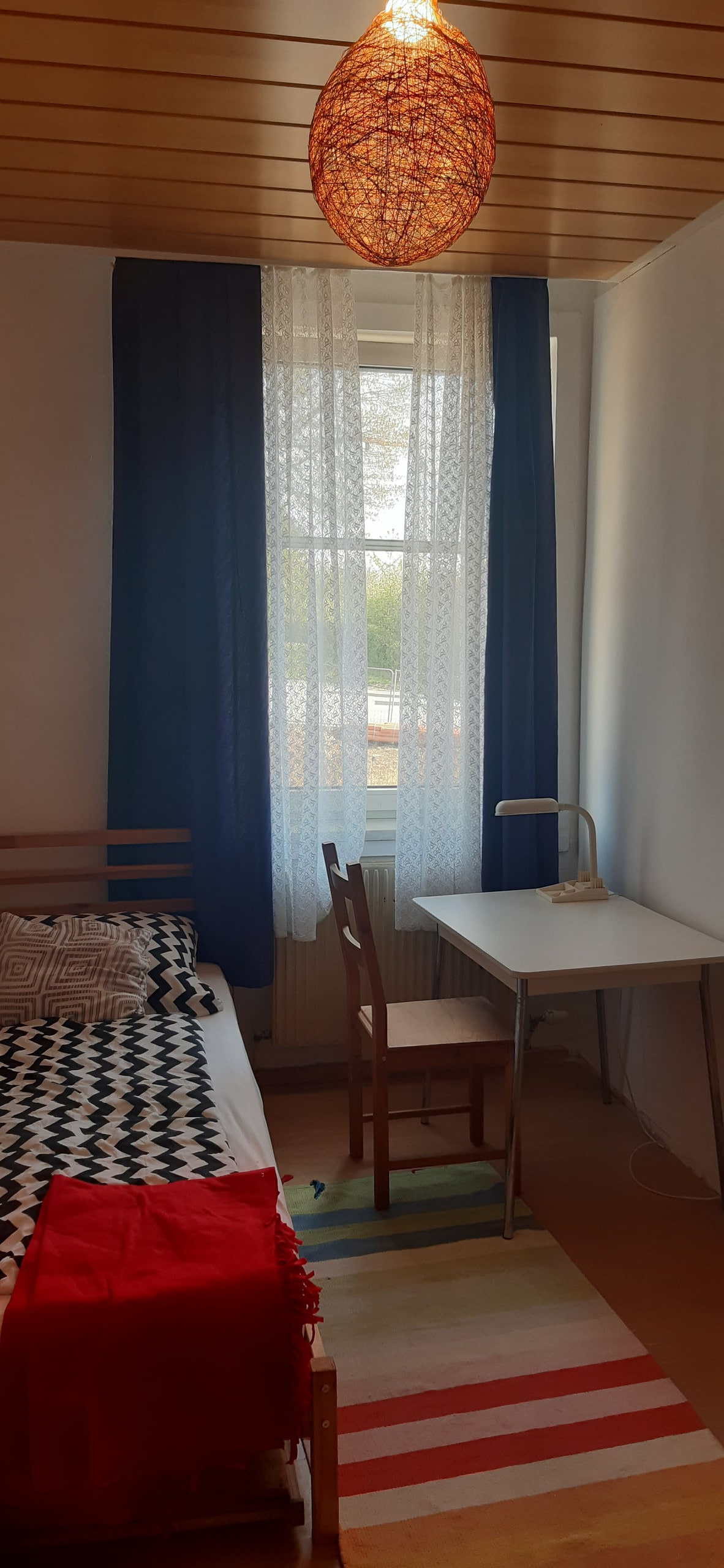 A cozy bedroom features a single bed with a patterned blanket, complemented by a simple desk and chair. Natural light enters through a large window dressed in lace and dark curtains. A colorful area rug adds a touch of vibrancy to the wooden floor.