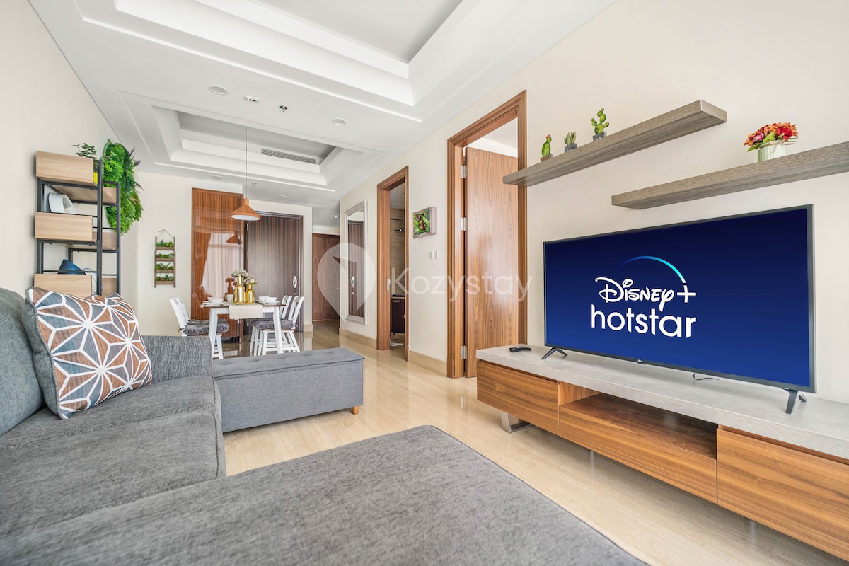 A contemporary living space features a gray sectional sofa adorned with patterned cushions. A wooden entertainment unit holds a large television displaying Disney+ and Hotstar. A dining area with a table for four and decorative plants adds a touch of vibrancy to the room.