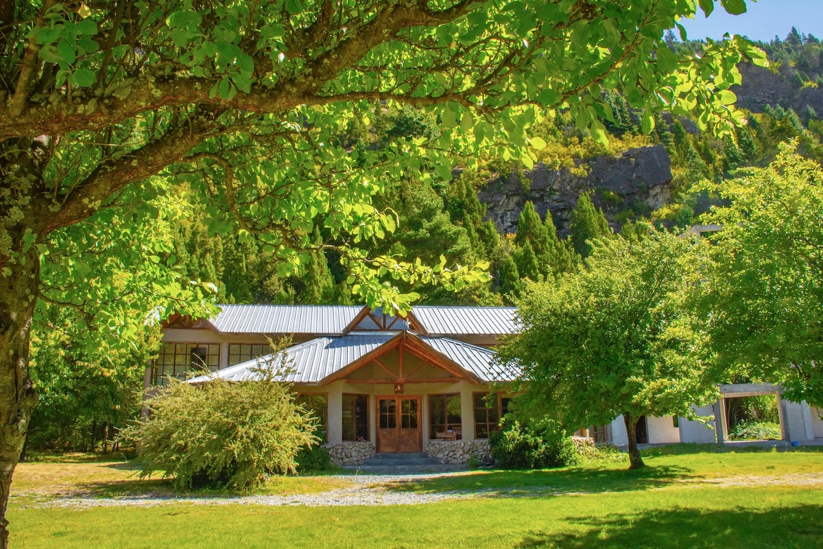 A spacious lodge is framed by lush greenery, featuring large windows that reflect the surrounding nature. The building's stone foundation and wooden beams provide a natural aesthetic, while the expansive lawn invites relaxation under the shade of several trees.