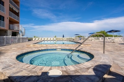 A well-maintained pool area features a hot tub alongside a spacious swimming pool, both surrounded by paved stone flooring. Beach towels are placed on lounge chairs under umbrellas, and a clear blue sky stretches overhead.