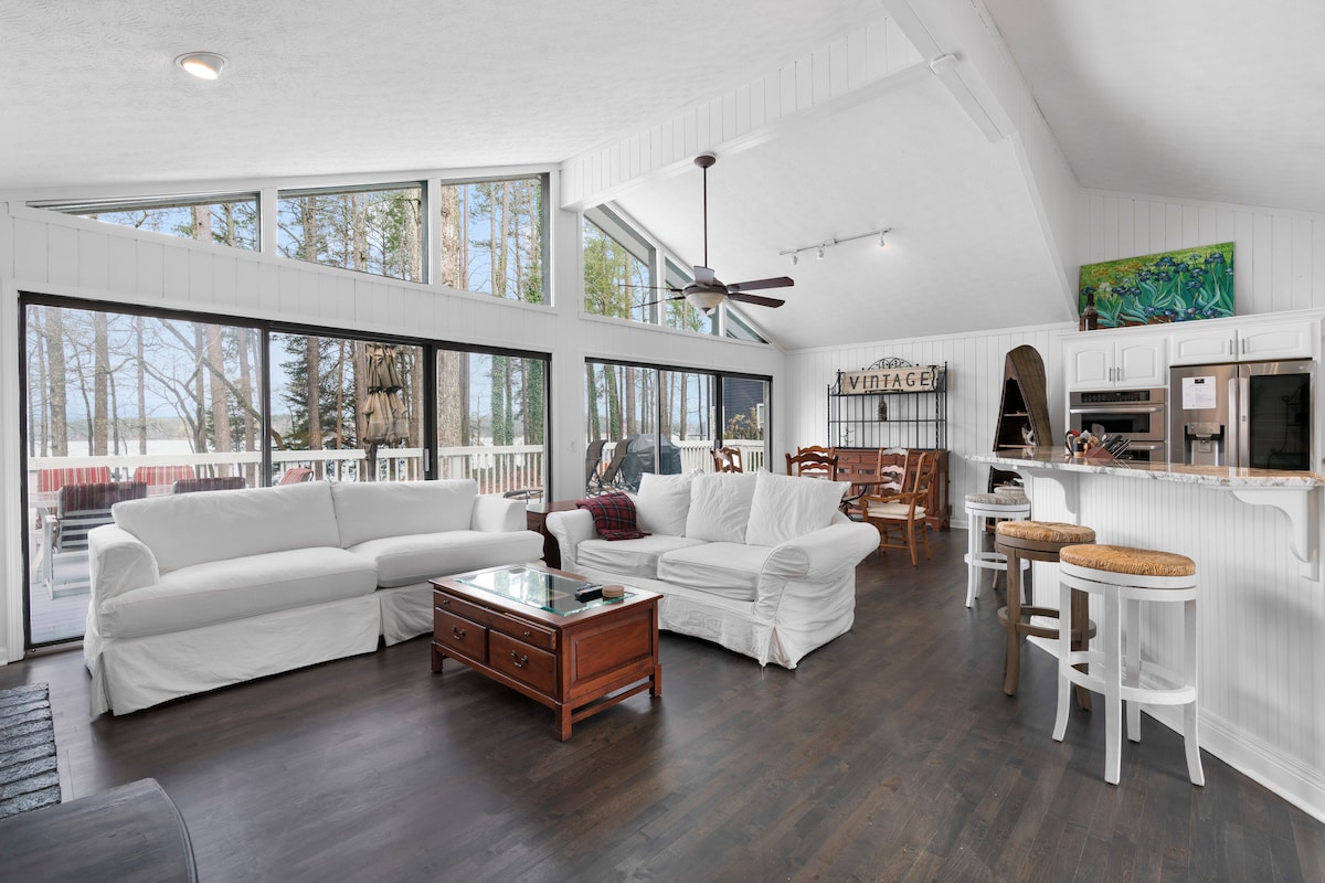 The living area features large windows that provide abundant natural light and views of the lake. Seating includes two slipcovered sofas and a wooden coffee table, with additional bar stools positioned at the kitchen counter. The open layout connects seamlessly with the dining space.
