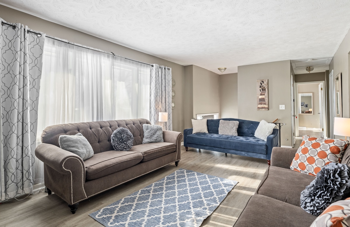 A spacious living area features a combination of two sofas, one in a rich blue and another in a warm brown. Soft throw pillows add comfort, while abundant natural light filters through large windows, enhancing the inviting atmosphere. A textured area rug lies on the floor.