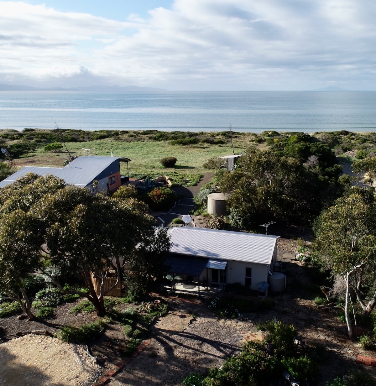 An aerial view captures the Dunes studio and its tranquil surroundings, framed by native trees. The glistening waters of the beach are visible in the background, along with lush greenery and a garden area, emphasizing the serene coastal setting.