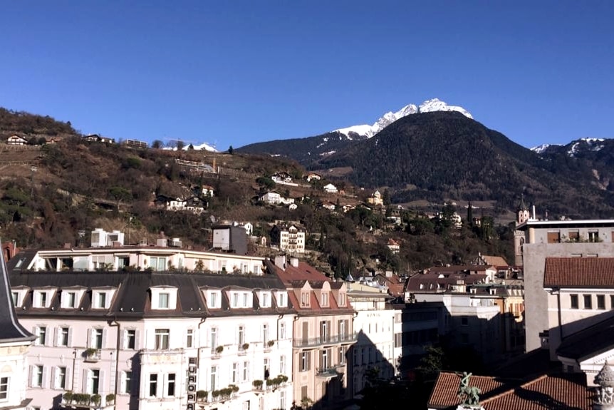 A panoramic view of a mountainous landscape is visible in the background, complemented by buildings of varying styles in the foreground. The sky is clear, showcasing bright blue hues, while the peaks are capped with snow.