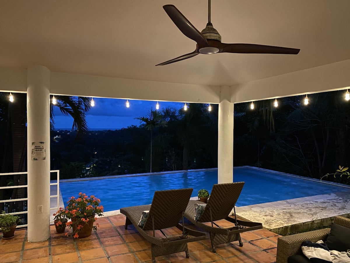 An outdoor terrace features two lounge chairs positioned near an infinity pool, which reflects the evening sky. Soft lighting creates a serene atmosphere, while lush greenery surrounds the area, enhancing the sense of privacy and tranquility.