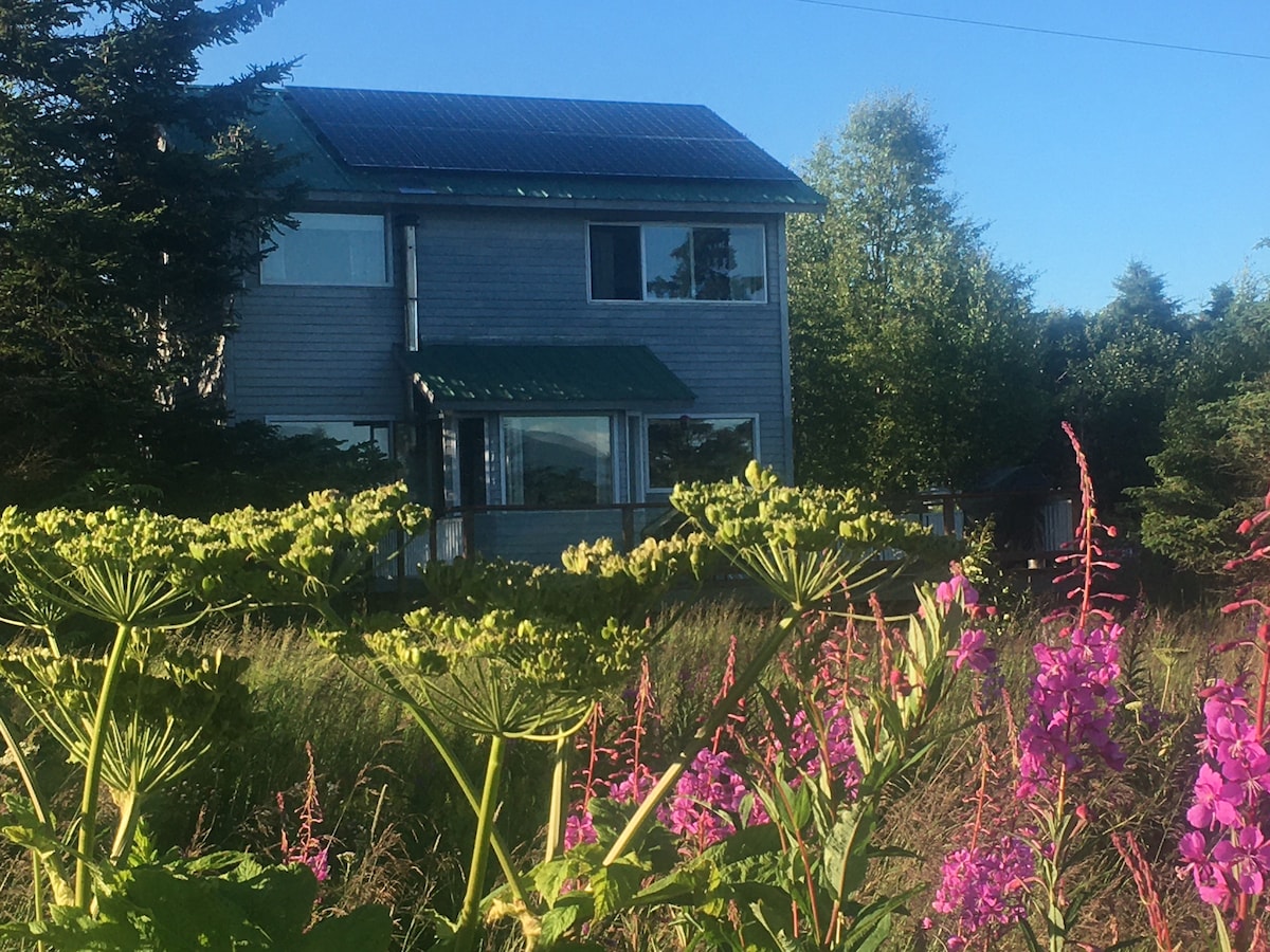 The exterior of a two-story blue bungalow is framed by colorful flowering plants, with pink and purple blossoms prominently visible in the foreground. A green metal roof complements the structure, which features large windows allowing natural light to penetrate the interior. Trees and shrubbery surround the property.