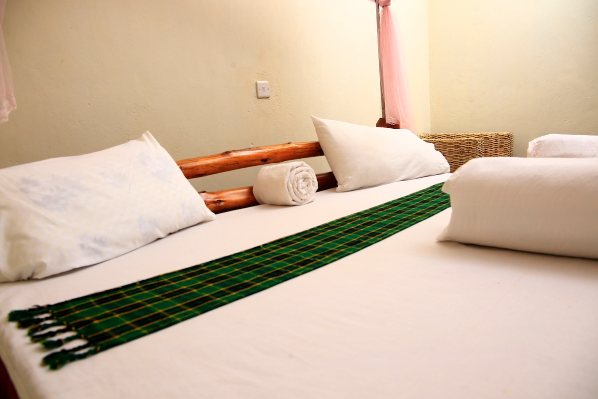 A cozy sleeping area features a spacious bed with locally made wooden accents. Two white pillows and rolled towels are placed neatly on the crisp white linens. A decorative green and black woven mat is displayed across the bed, adding a touch of local craftsmanship.