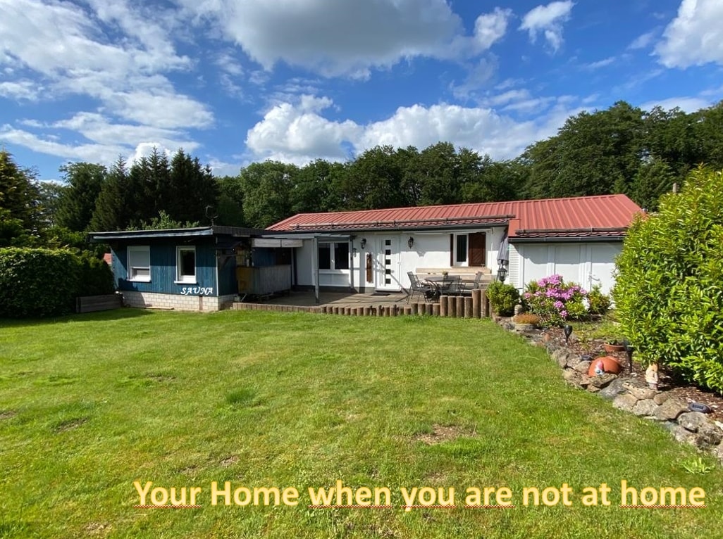 A charming exterior view of the accommodation is displayed, featuring a lawn surrounded by greenery and flower beds. The building features a combination of a red metal roof and light-colored walls, with patio seating visible in front. Blue shutters adorn the windows.