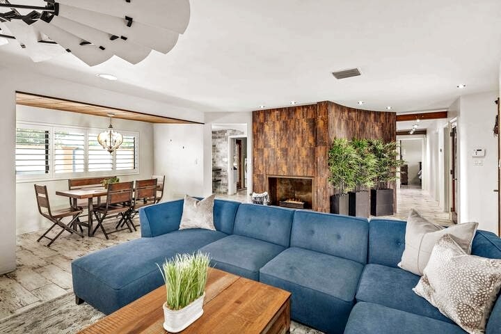 A spacious living area features a blue sectional sofa complemented by a wooden coffee table. Natural light enters through the nearby windows, illuminating the contemporary decor. A dining area with wooden chairs and a decorative light fixture is visible in the background, enhancing the inviting atmosphere.