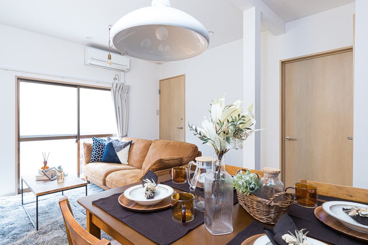 A dining area features a polished wooden table set with plates and glasses, complemented by a woven basket. A comfortable couch is situated nearby, with decorative pillows enhancing the space. Natural light flows in through large windows, illuminating the warm color palette.