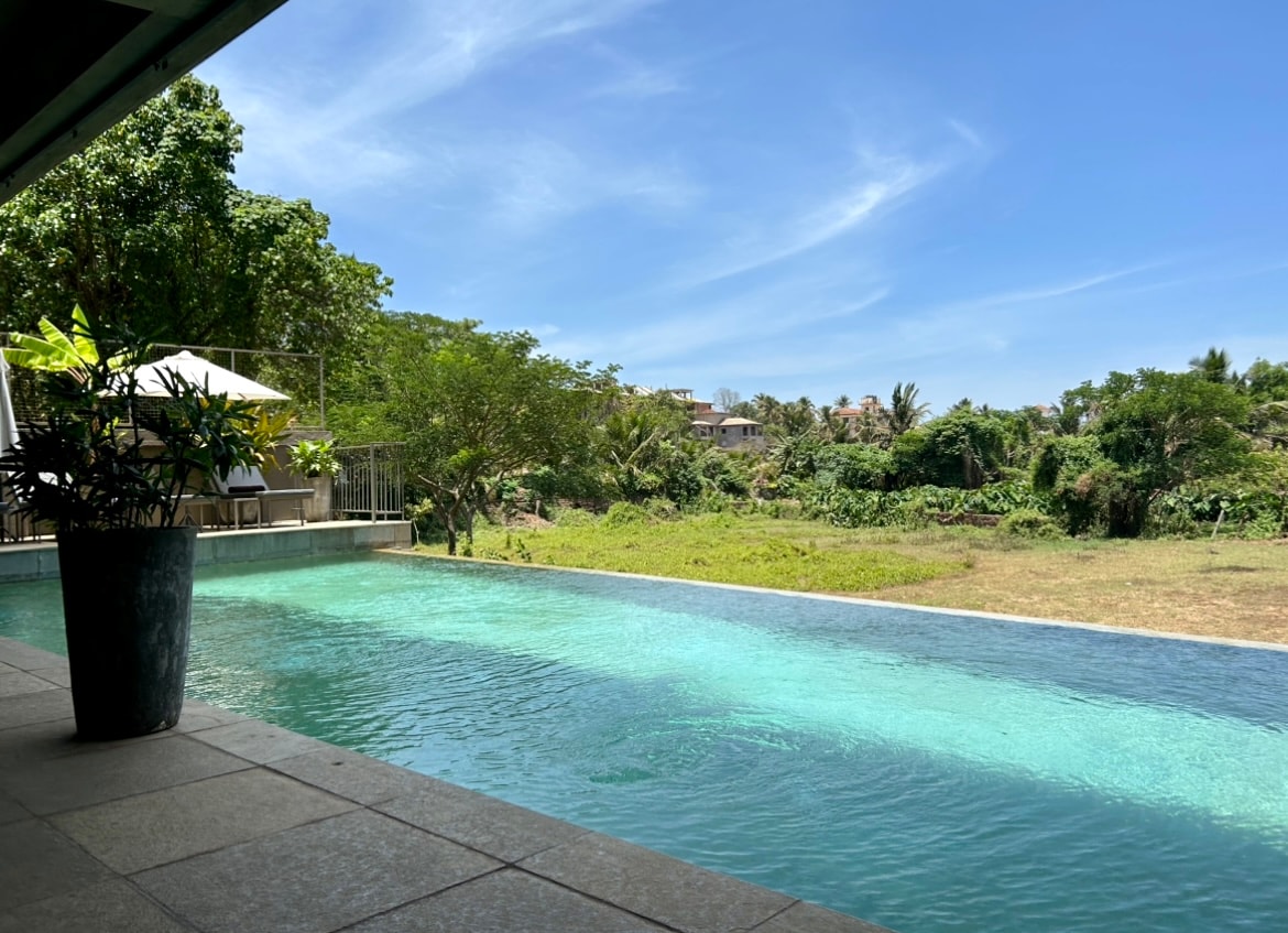 An infinity pool extends along the outdoor lounge area, reflecting the clear blue sky. Lush greenery surrounds the pool, offering a serene view of the landscape. Sunlight filters through the trees, enhancing the tranquil atmosphere of the space.
