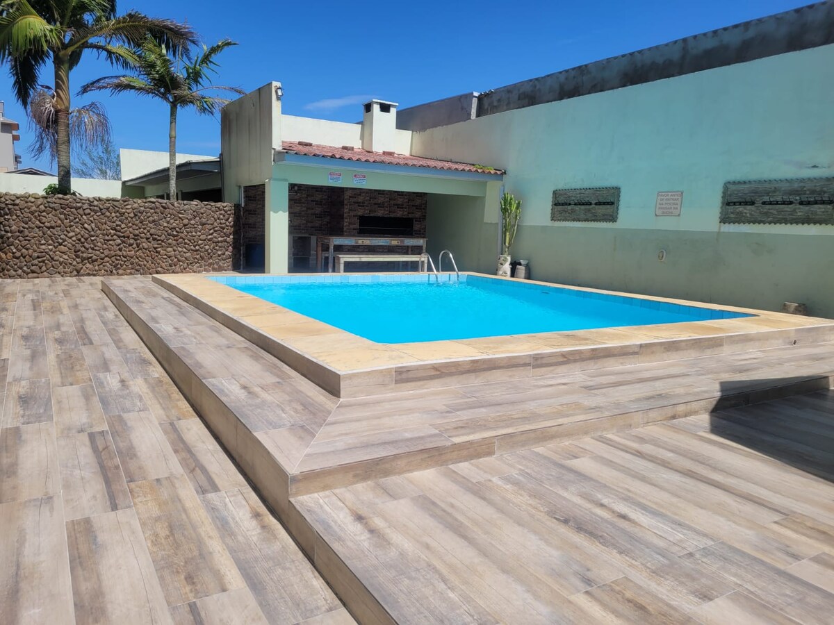 A rectangular swimming pool featuring clear blue water is surrounded by light-colored tiled decking. A covered outdoor area with a grill is visible in the background, complemented by a stone wall and palm trees that add a natural touch to the setting.