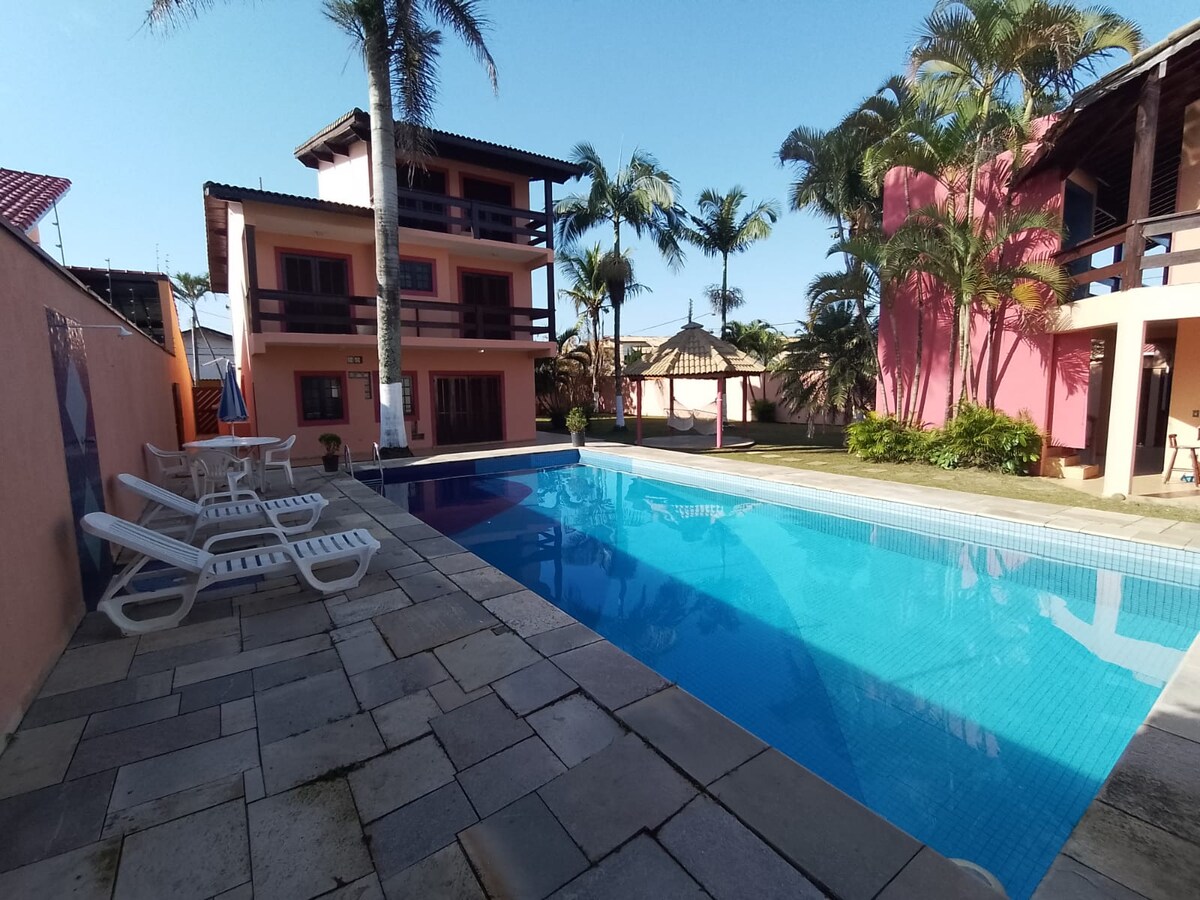 A large swimming pool is framed by palm trees and spacious lounging areas. Sun loungers are positioned beside the pool, set on a stone patio. Two multi-story buildings are visible in warm colors, creating a relaxed outdoor ambiance in the backyard.