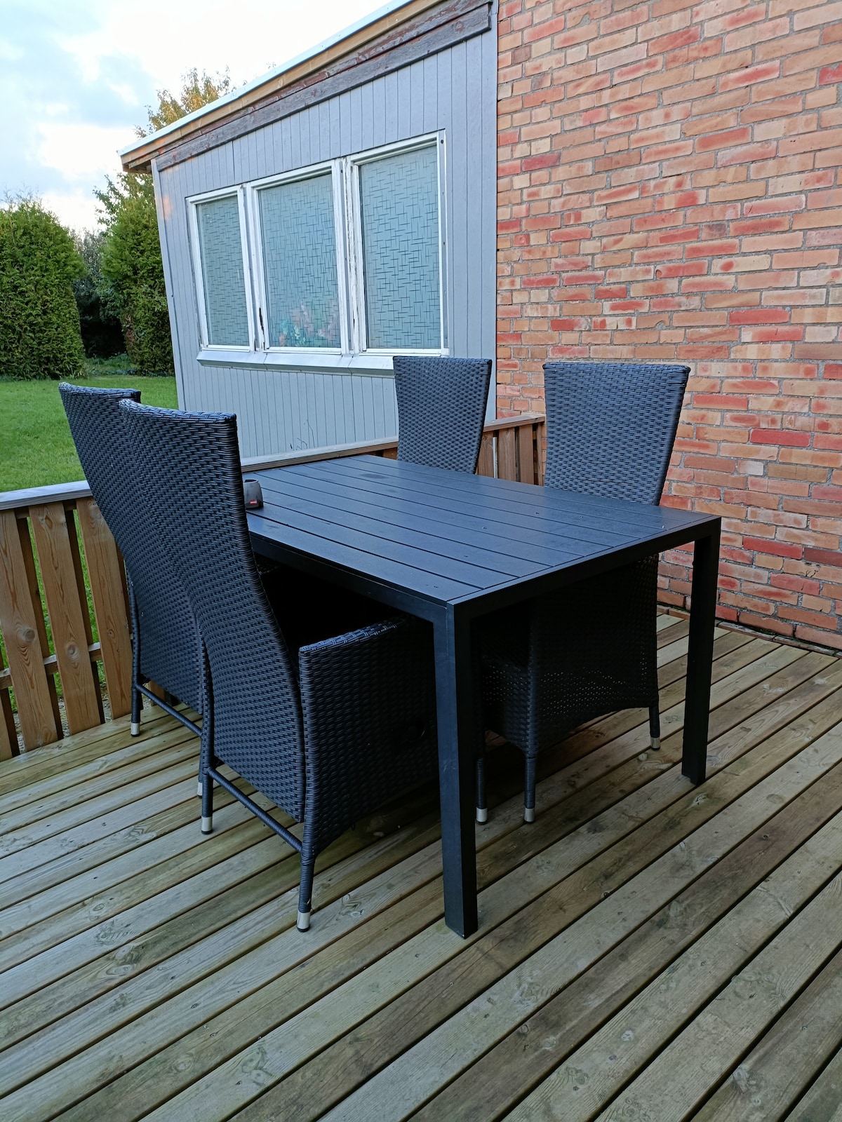 A dining set is positioned on a wooden deck, featuring a rectangular table surrounded by four woven chairs. The area is enclosed by a wooden railing, with green grass visible in the background and large windows of a building visible.