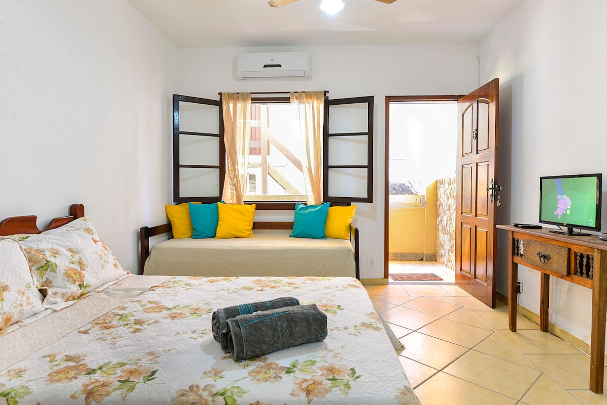 A well-lit bedroom features a spacious bed adorned with a floral-patterned blanket. A cozy seating area is visible, highlighted by bright yellow cushions. Air conditioning is available, and a TV provides entertainment. Doorways lead to an adjoining space, enhancing the room's accessibility.
