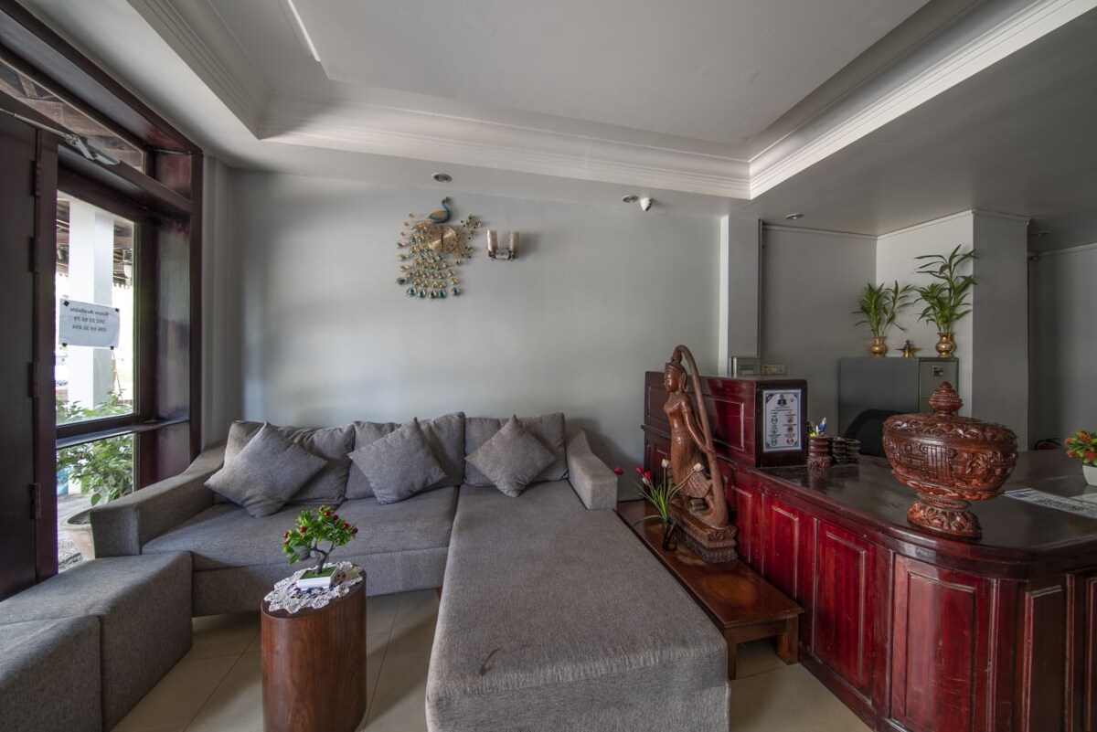 A spacious reception area features a large gray sectional sofa alongside a wooden reception desk. Decorative elements include a carved statue and a peacock wall hanging. Natural light enters through windows, enhancing the welcoming atmosphere of the space.