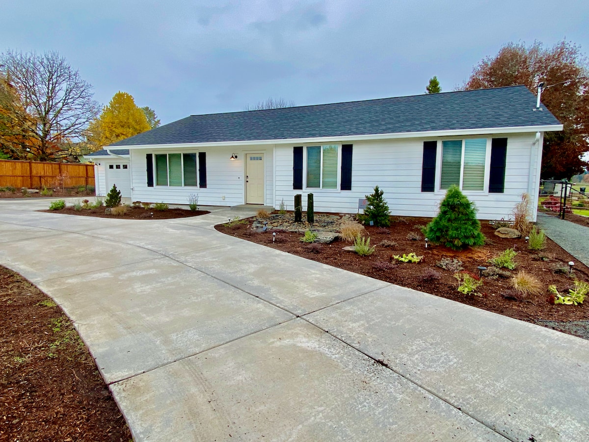 A single-level home is viewed from a distance, featuring a large driveway and well-maintained landscaping. Modern windows with dark shutters are visible, along with a symmetrical entryway. The surrounding yard includes small shrubs and patches of grass, contributing to an inviting exterior.