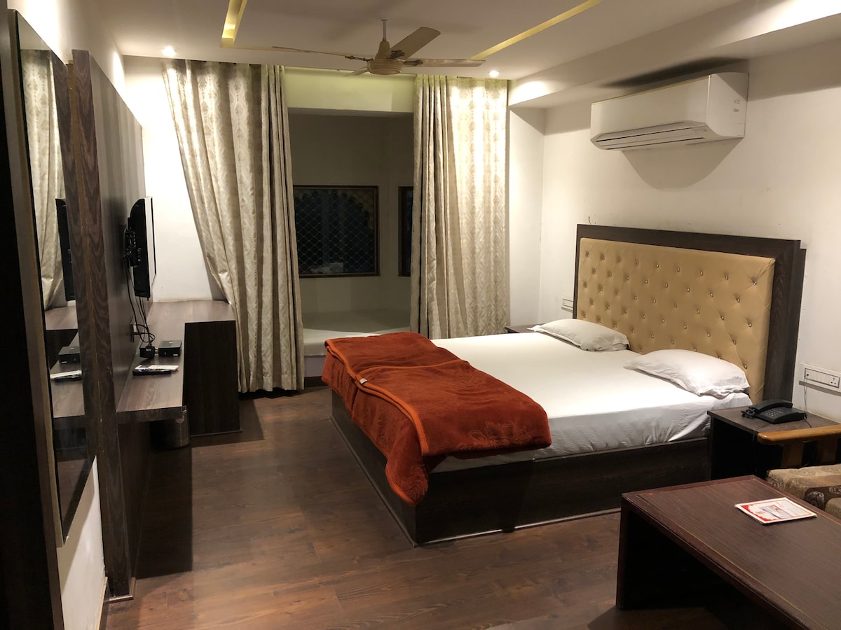 A spacious bedroom features a large bed adorned with a soft orange blanket. Light-colored walls enhance the brightness, while dark wood furnishings provide contrast. Curtains frame the windows, and a flat-screen TV and desk are visible, contributing to the modern decor.