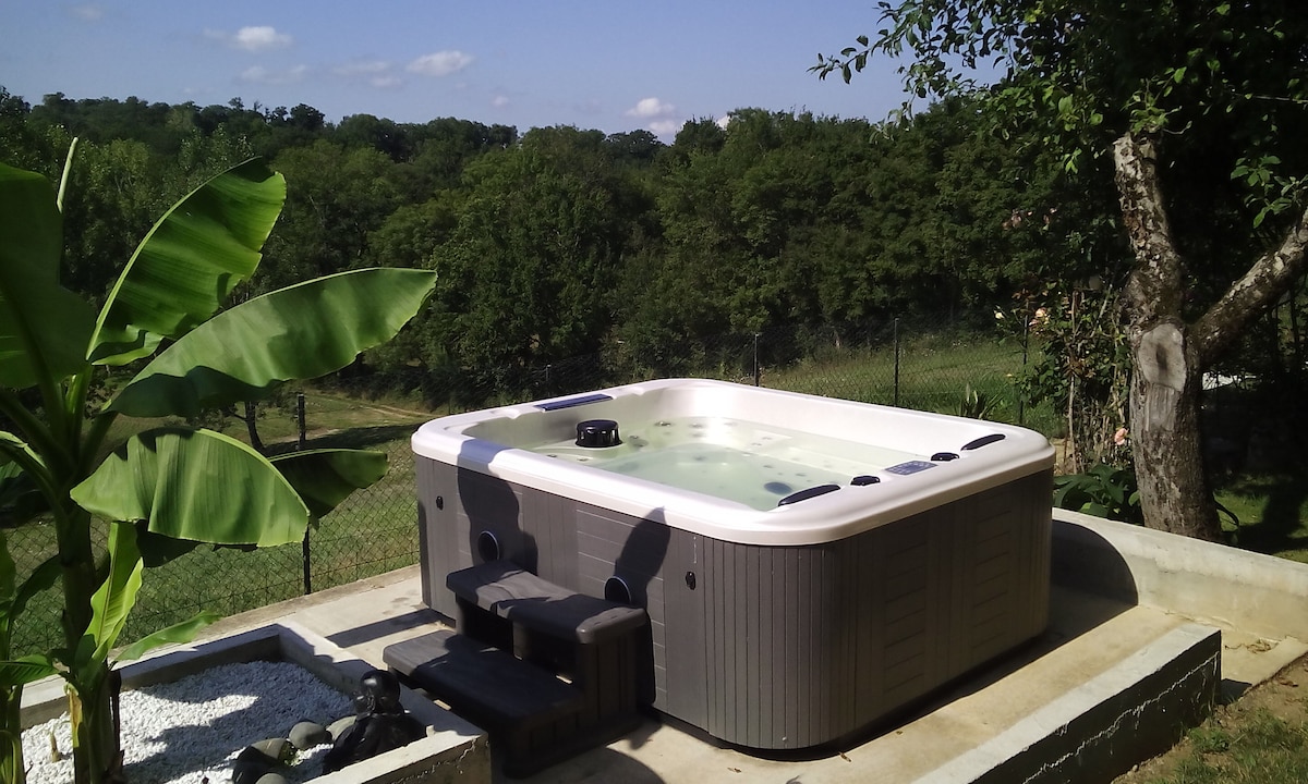 A private jacuzzi sits on a concrete patio, surrounded by lush greenery. A banana plant is visible nearby, and the landscape features rolling hills in the background. The calm water reflects the clear blue sky, creating a serene outdoor space.