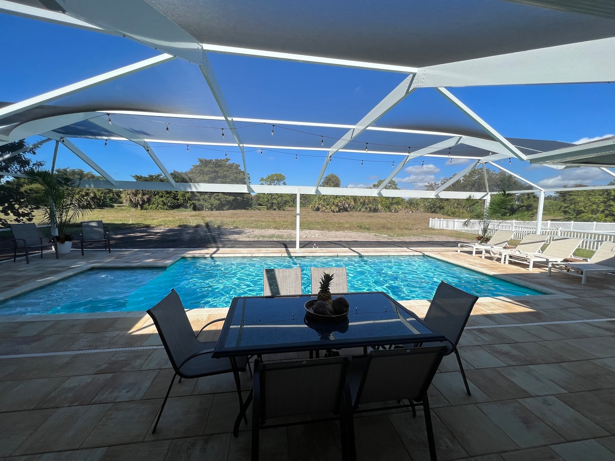 A spacious pool area is enclosed by a caged structure, featuring a heated swimming pool. A dining table is set with seating for six, complemented by lounge chairs around the pool. Lush landscaping is visible behind the pool area, under a clear sky.