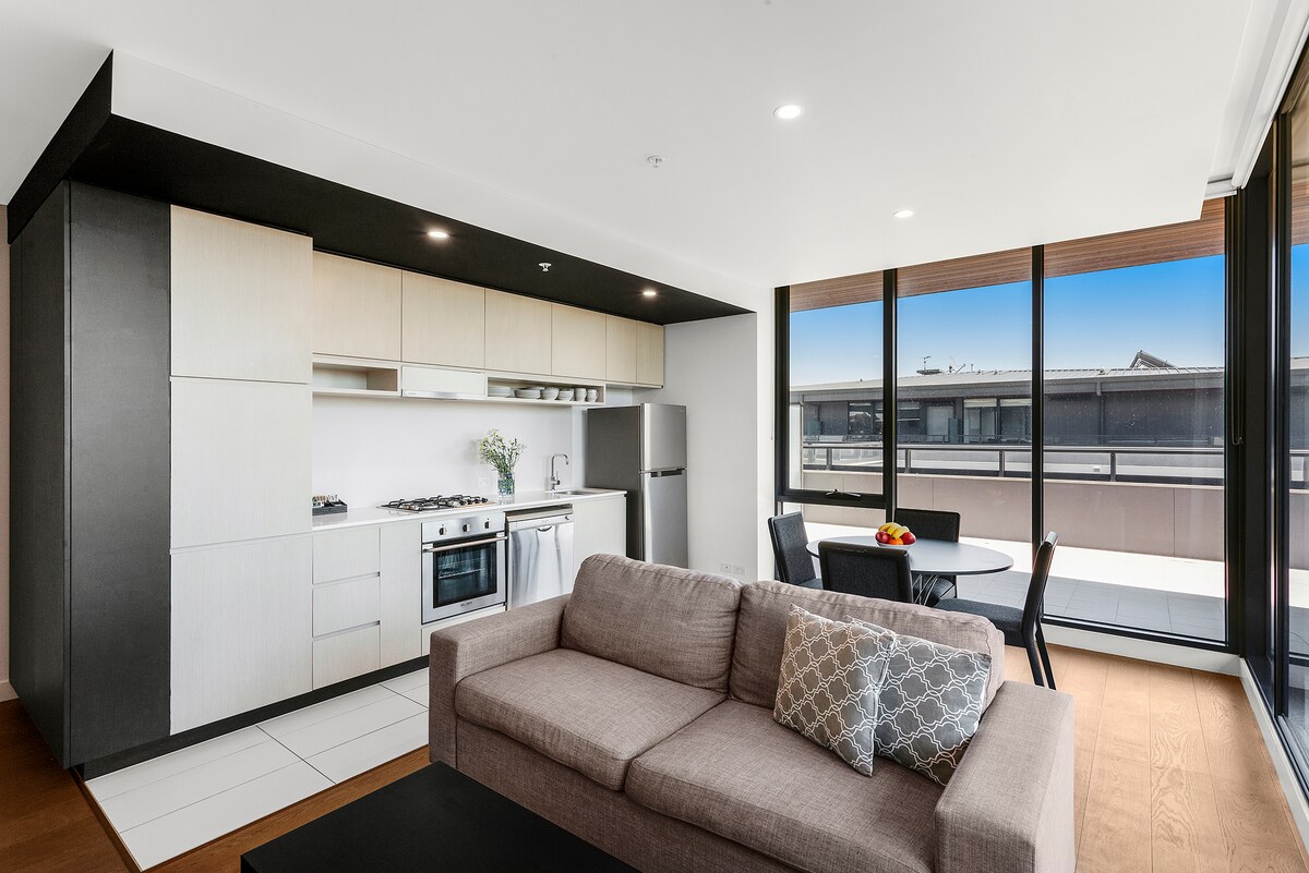 A modern living area includes a comfortable sofa, dining table, and fully equipped kitchen featuring stainless steel appliances. Large windows offer ample natural light and views of the private balcony. The sleek design is characterized by clean lines and a neutral color palette.