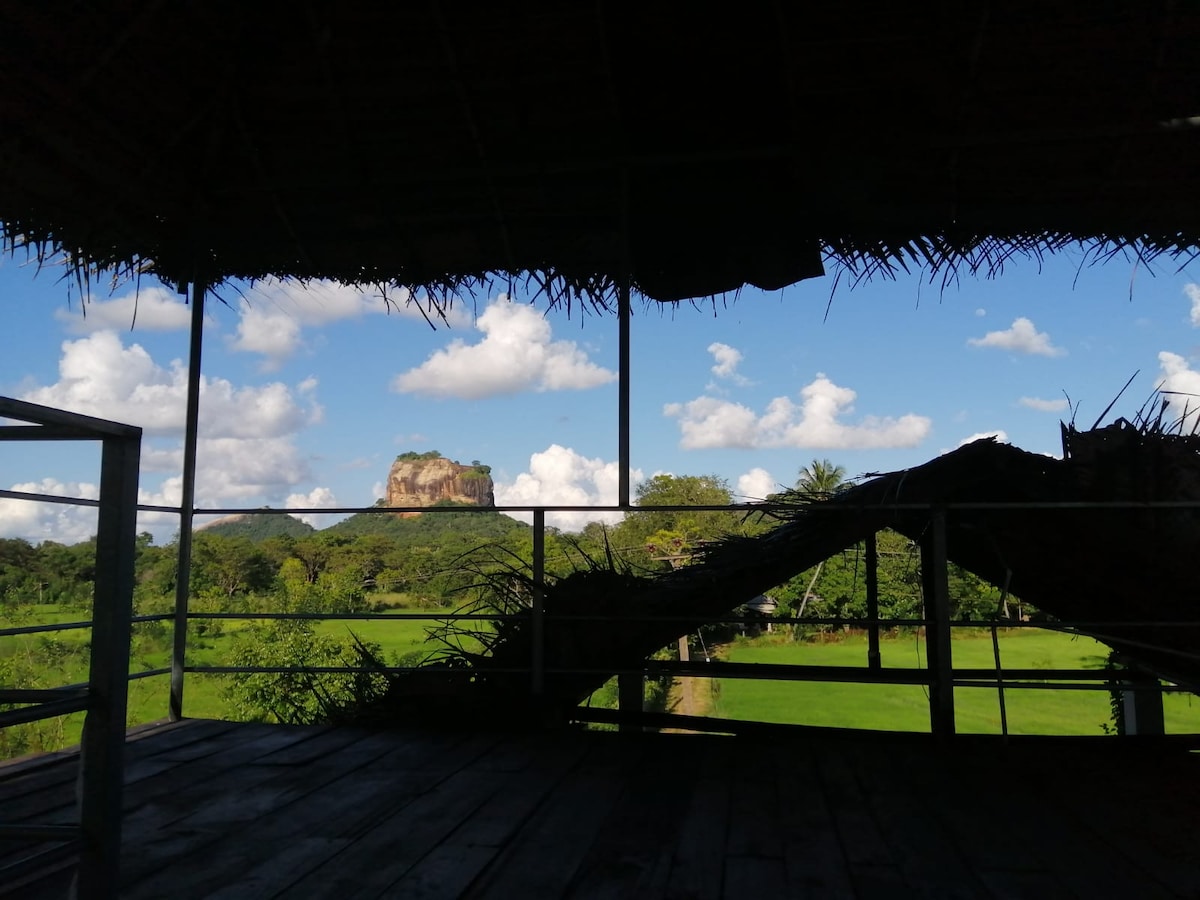 The view from the open balcony frames Sigiriya Lion Rock, visible amidst a backdrop of lush greenery and vibrant blue skies adorned with scattered clouds. The natural landscape emphasizes the tranquil setting, creating a serene escape in the countryside.
