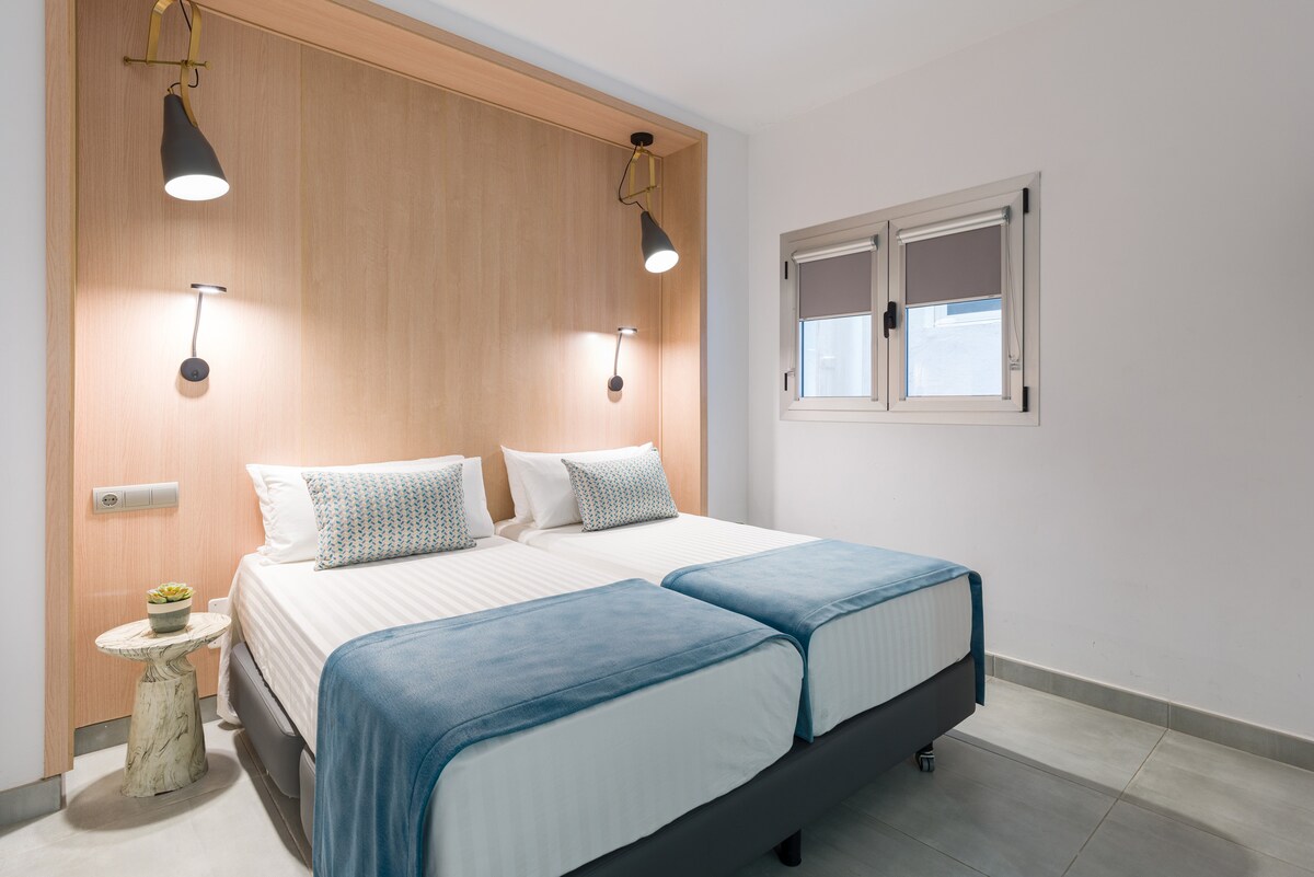 A modern room features two single beds with light linens and a soft blue throw. A minimalist side table is positioned beside one bed, while wall-mounted lamps provide illumination. An adjustable window with blinds allows natural light to enter the space.
