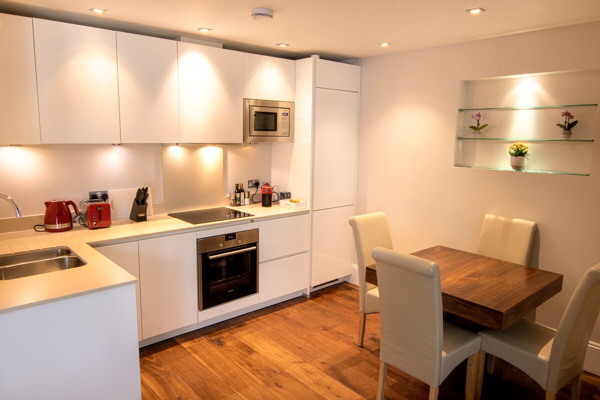 A modern kitchen is equipped with sleek white cabinetry and stainless steel appliances. A wooden dining table is surrounded by four upholstered chairs. Soft lighting highlights the countertops, while a decorative shelf displays a small potted plant near a window.