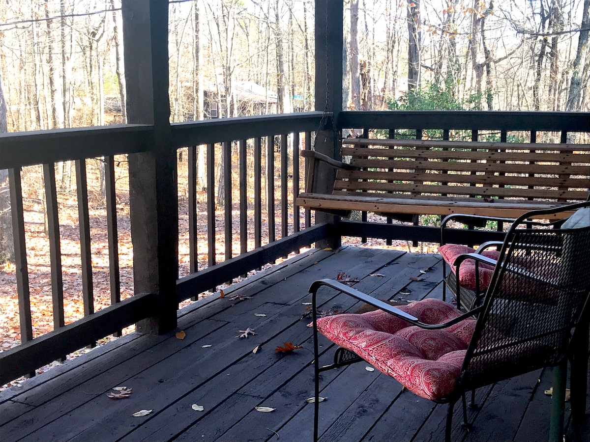 A spacious porch features a wooden swing and two metal chairs, providing a comfortable spot to enjoy the surrounding woods. Fallen leaves scatter the dark wooden floor, while the railing offers a view of the natural setting outside.