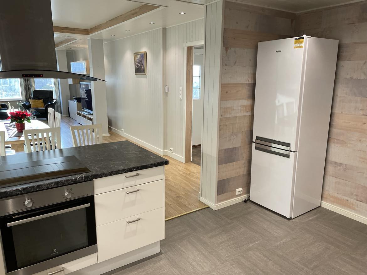 A modern kitchen features a sleek black countertop and contemporary cabinetry. A stainless steel stove and hood are prominently displayed, while a spacious white refrigerator is located to the side. Bright light fills the area, enhancing the open layout that connects to the living space.