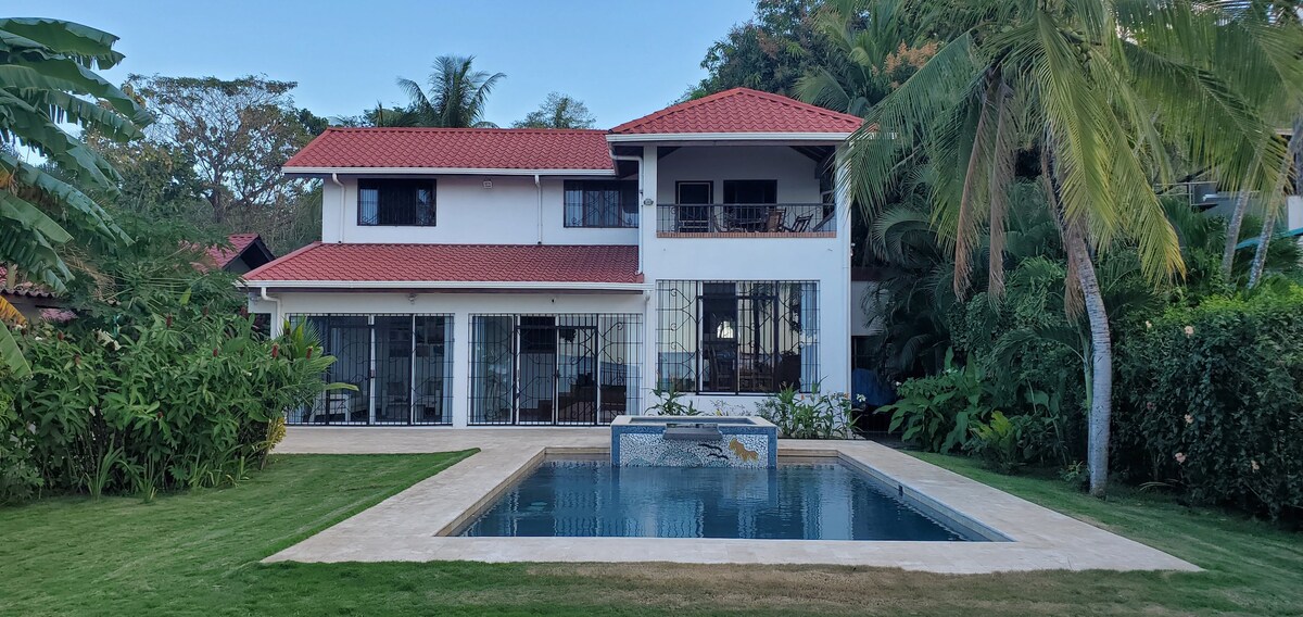 The exterior view captures a two-story home surrounded by lush greenery. The vibrant red roof contrasts with the white walls. A private pool is positioned in the foreground, and covered balconies are visible on the upper level, enhancing the outdoor living space.