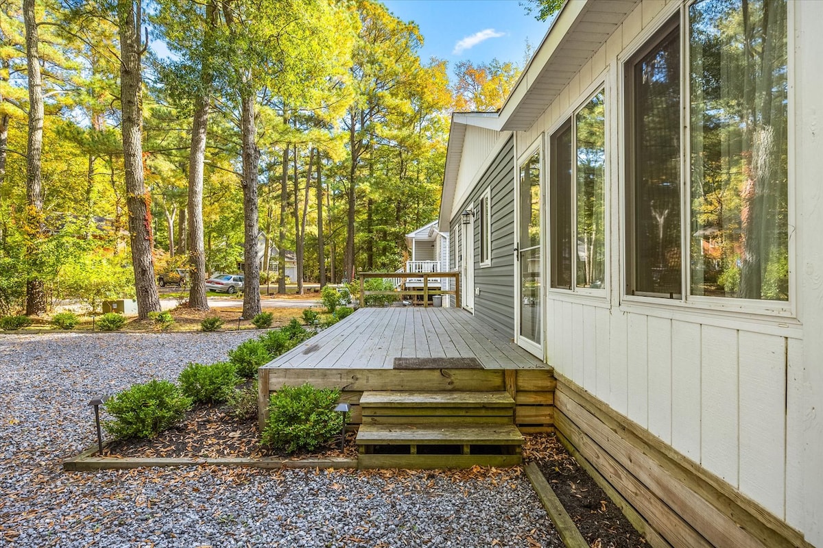 A wide wooden deck is visible, leading to the entrance of the house. Surrounding greenery includes shrubs and trees, providing a natural setting. The pathway is lined with gravel, while large windows allow for ample natural light inside.