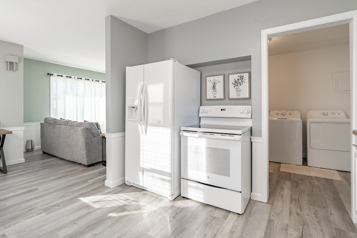 A well-lit kitchen area features modern white appliances, including a refrigerator and electric stove. The spacious layout includes access to a laundry room through an open doorway, with sunlight enhancing the room's neutral color palette and adjoining comfortable living space.