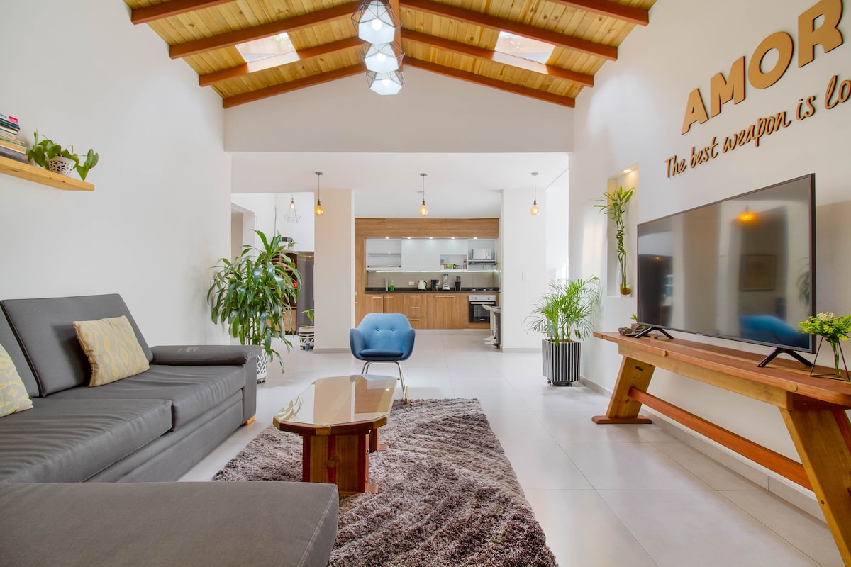 A spacious living area features a cozy gray sofa with bright cushions and a sleek coffee table atop a plush rug. A modern blue accent chair adds color, while indoor plants bring greenery. The kitchen is visible in the background, enhancing the open layout.