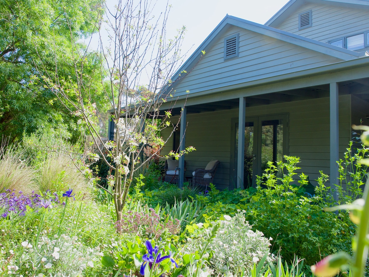 A serene exterior view showcases the entrance of a residence surrounded by lush gardens. Various plants and colorful flowers fill the landscape, while a small tree stands nearby. Two chairs are positioned on the porch, inviting relaxation under natural light.